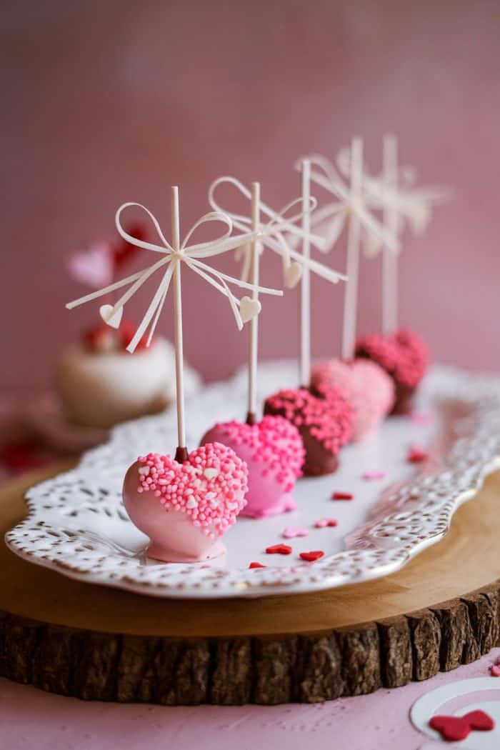 Heart-shaped cake pops coated in pink and red sprinkles are arranged on a decorative white platter. Each pop is adorned with a stick tied with a bow. The background features soft, warm tones of pink.