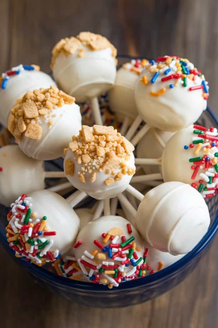 A bowl filled with white chocolate cake pops. Some are decorated with colorful sprinkles, while others are topped with crushed cookie bits. Each cake pop is on a stick, and they are arranged in a cluster, creating a festive appearance.