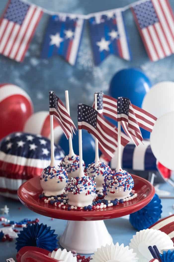 Cake pops decorated with red, white, and blue sprinkles and small American flags are displayed on a red platter. The background features patriotic balloons and bunting in stars and stripes, creating a festive atmosphere.