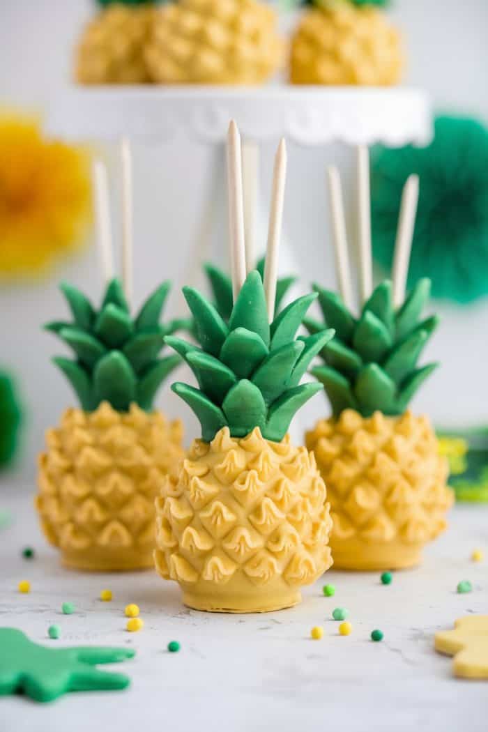 Four cake pops are decorated to look like pineapples, complete with leafy green tops and textured yellow bodies. They are presented on wooden sticks with a white cake stand and colorful decorations as a backdrop.