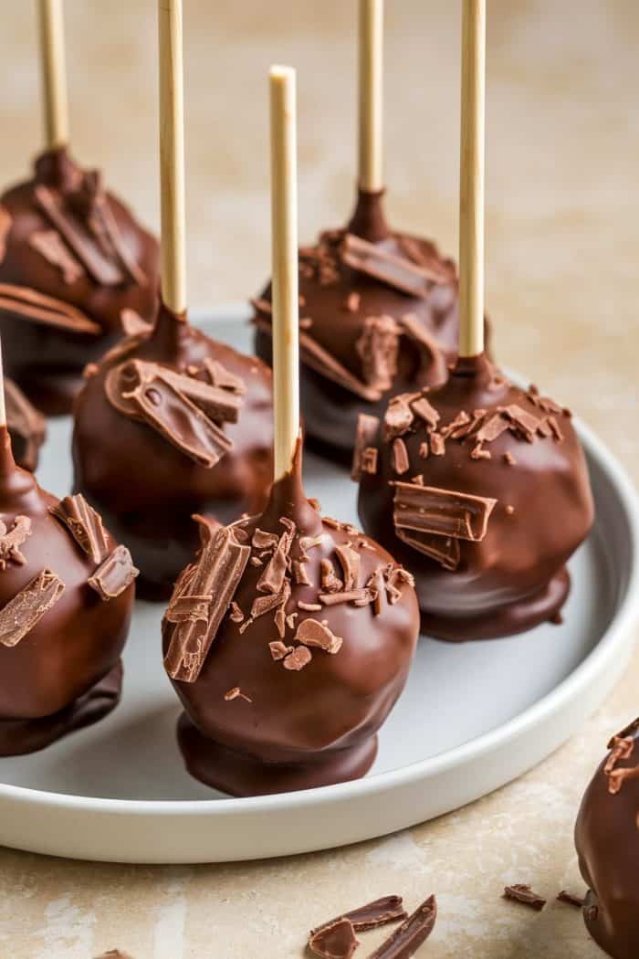 Chocolate cake pops arranged on a white plate, each with wooden sticks and topped with chocolate shavings. The background is a light surface, enhancing the appetizing display.
