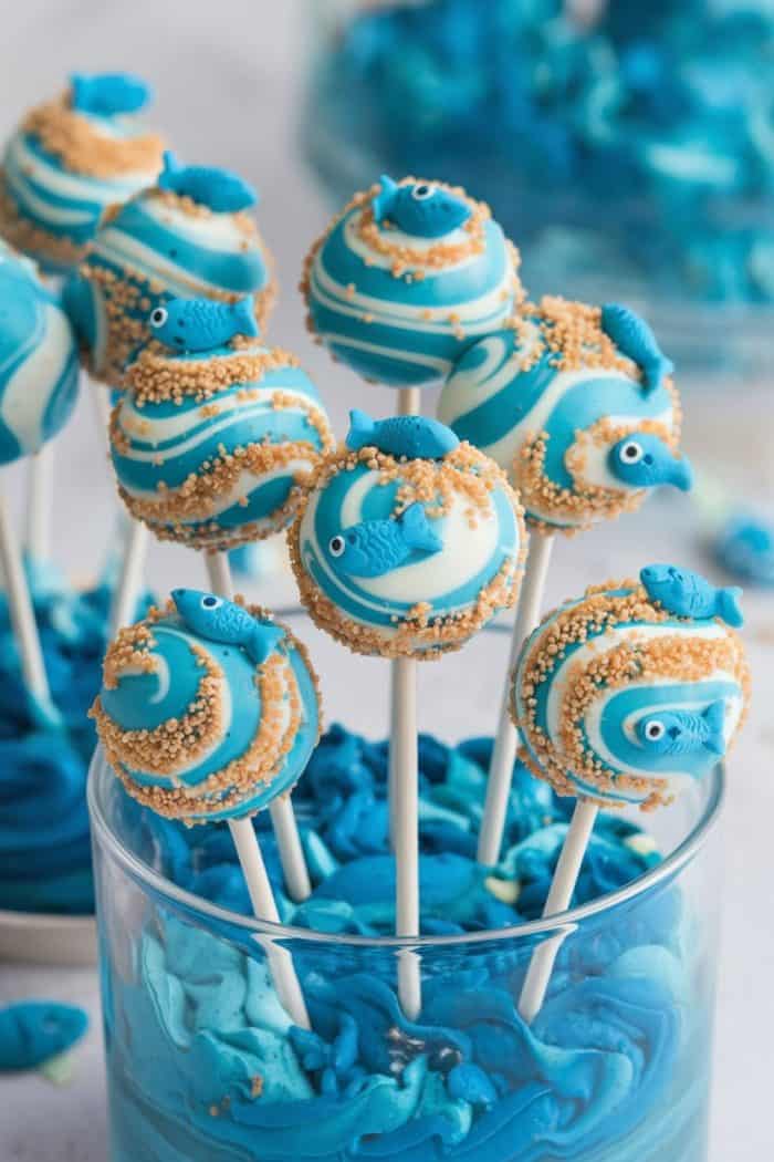 Round cake pops decorated with swirled blue and white icing, topped with small blue fish-shaped candies and sprinkled with cookie crumbs. They are displayed in a glass filled with blue frosting for a sea-themed presentation.