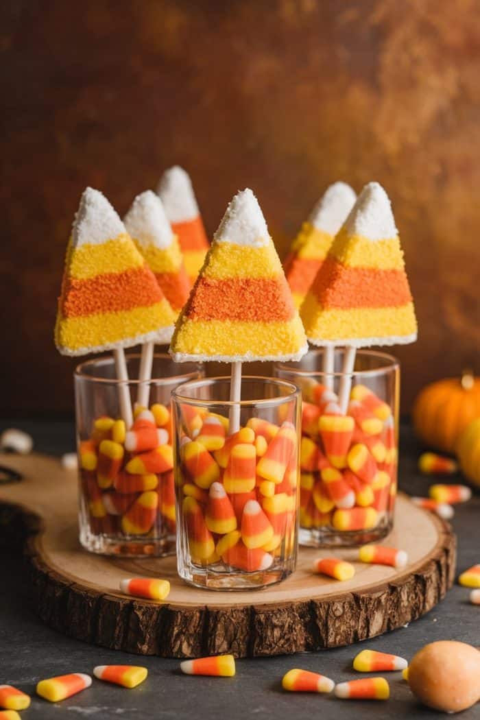Four triangular cake pops decorated to look like candy corn stand upright in glasses filled with candy corn, set on a wooden slab. Additional candy corn pieces are scattered on a dark table, with a small pumpkin in the background.