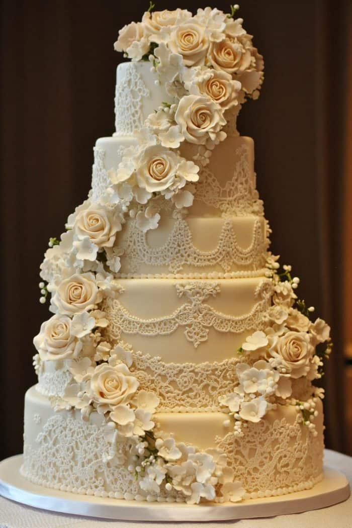 A tall, elegant wedding cake with multiple tiers, decorated with intricate white lace patterns and cascading white roses. The floral decorations add a touch of sophistication, creating a luxurious and romantic appearance.