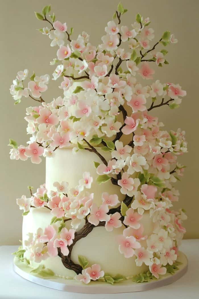 A two-tiered cake adorned with realistic pink and white cherry blossoms cascading down the sides. The intricate sugar flowers and green leaves create a delicate, elegant design against a cream-colored background.