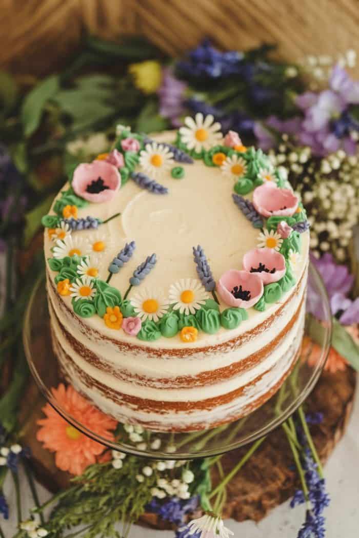 A rustic naked cake on a glass stand, decorated with buttercream flowers including daisies, poppies, and lavender. Surrounding the cake are various colorful wildflowers on a wooden surface.