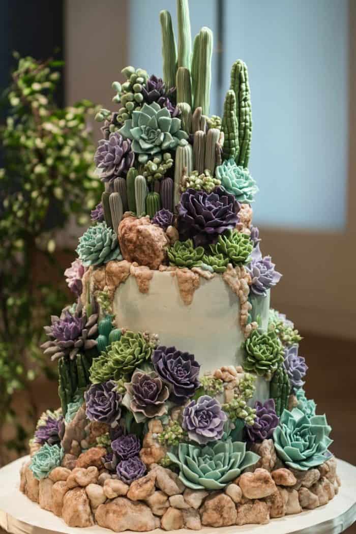 A tiered cake decorated with realistic succulents and cacti made from icing. The design includes various sizes and colors, such as green, purple, and teal, with sugar rocks at the base, creating a desert garden look.