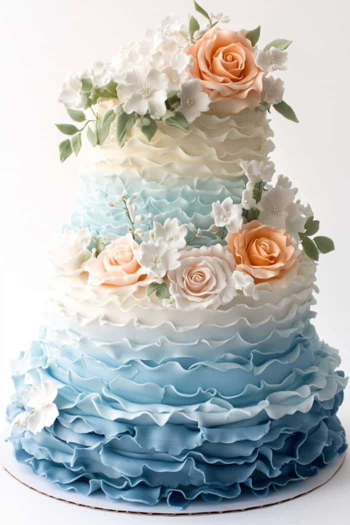 A tiered cake with a gradient ruffle design transitioning from white at the top to blue at the bottom. The cake is adorned with peach and white flowers, including roses and blossoms, along with green leaves.