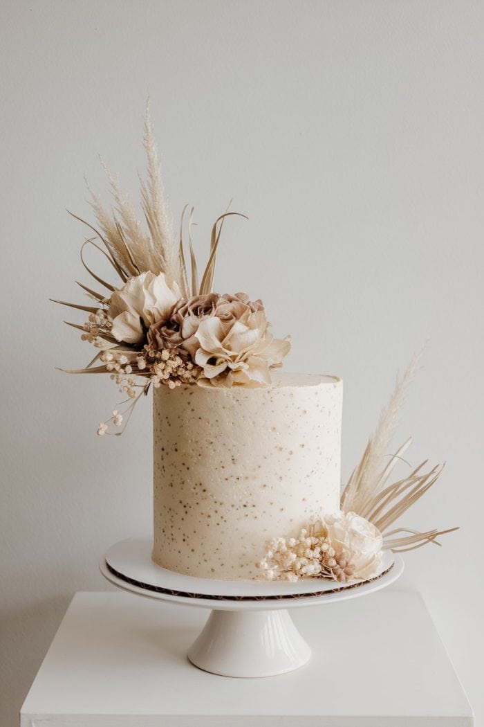 A tall, elegant cake with light beige frosting is adorned with dried flowers and foliage. It stands on a white cake stand against a neutral background.