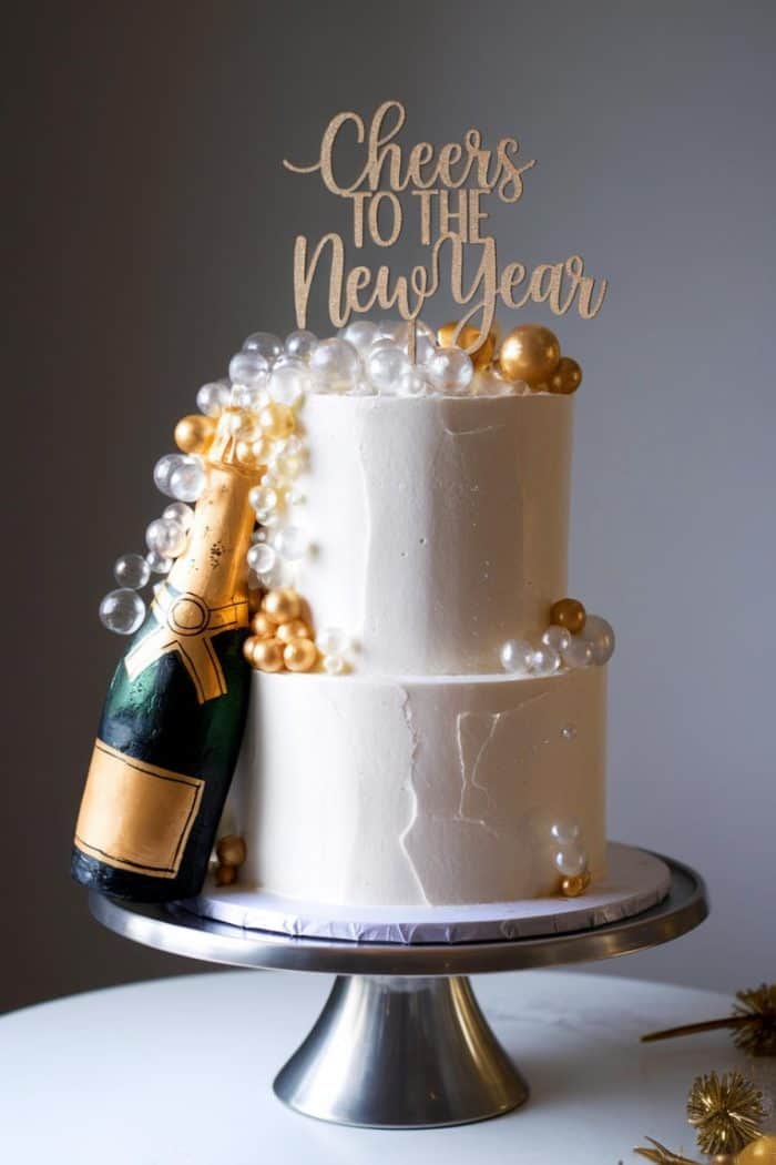 Two-tier white cake with gold and clear edible bubbles, adorned with a champagne bottle decoration. A topper reads "Cheers to the New Year." The cake is displayed on a silver cake stand.