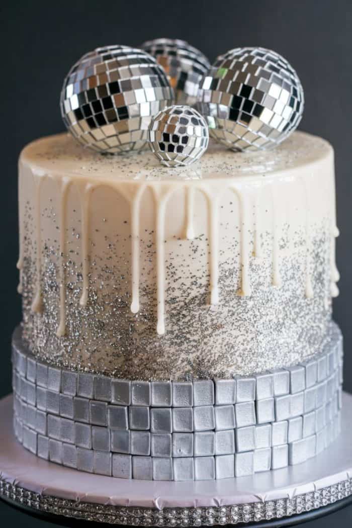 A cake with silver and white decorations features a smooth white frosting with a dripping effect. Silver glitter covers the middle section, and silver disco ball decorations top the cake. A silver tile pattern decorates the base.