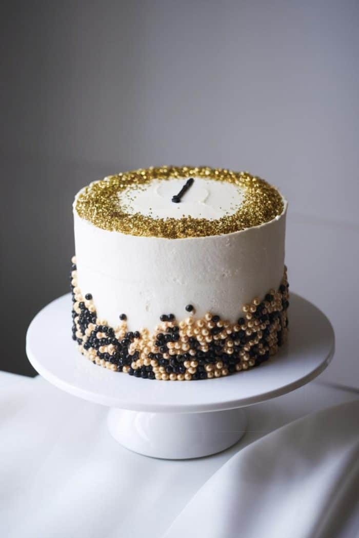 A white frosted cake on a stand, decorated with gold and black beads around the base and a ring of gold sprinkles on top. A single black candle is placed in the center.