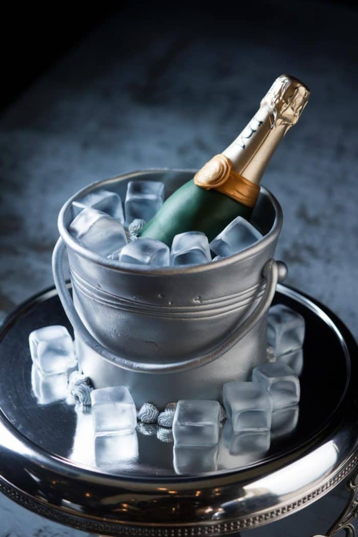 A cake designed to look like a champagne bottle in an ice bucket. The cake is detailed with metallic silver icing and ice cubes, sitting on a reflective silver tray.