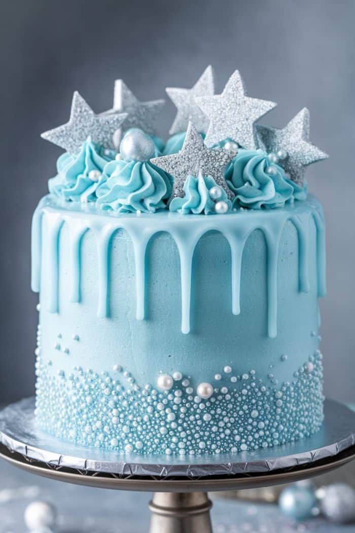 A blue cake with light blue frosting drips, decorated with silver star toppers, blue and silver spherical candies, and textured with small edible pearls, sits on a metallic cake stand against a grey background.