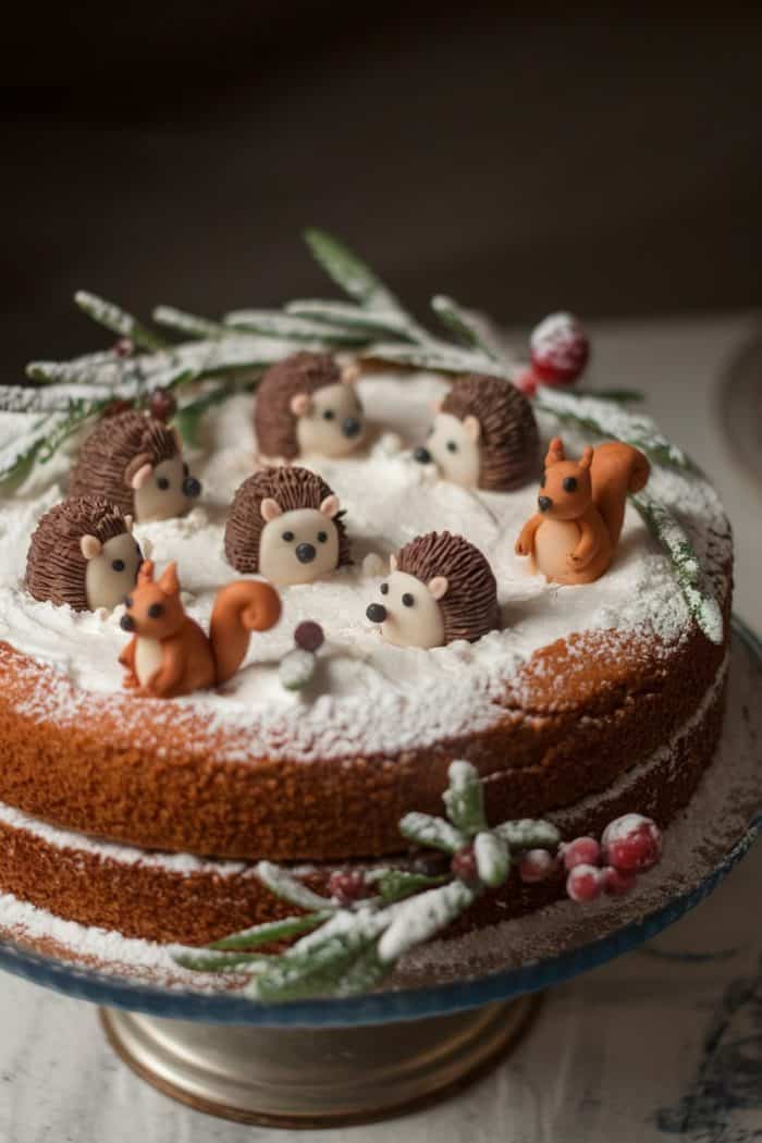 A cake topped with white icing and decorated with small, edible figures of hedgehogs and squirrels. It is garnished with powdered sugar, resembling snow, and accented with green leaves and red berries.