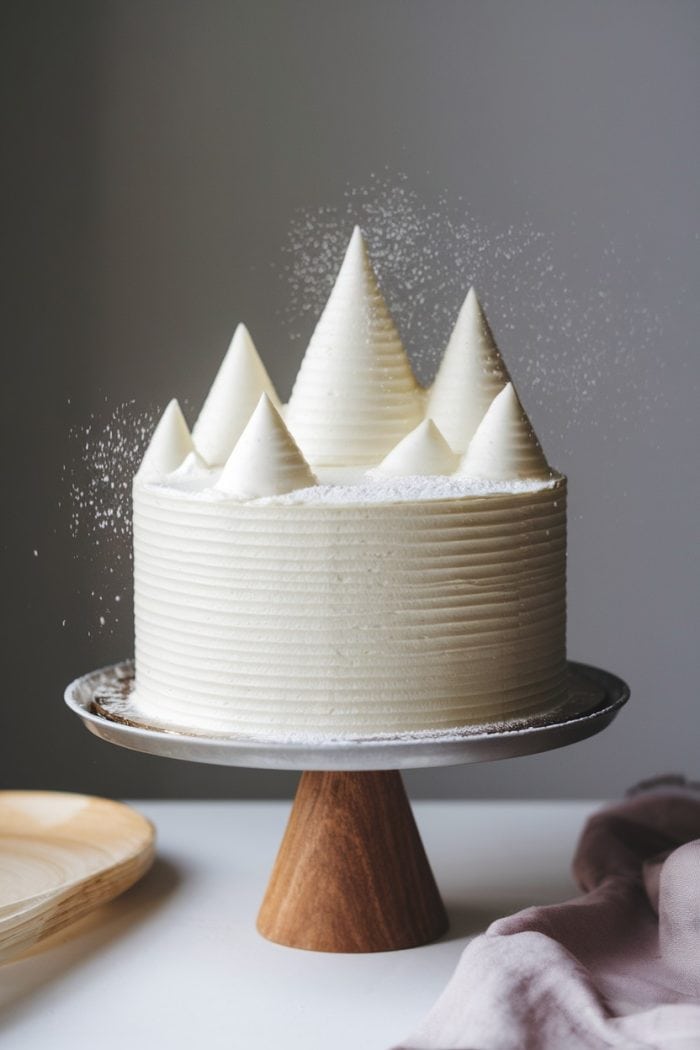 A white frosted cake with tall, pointed peaks resembling mountains, lightly dusted with powdered sugar. The cake sits on a wooden stand, and there is a wooden plate and a pink cloth nearby.