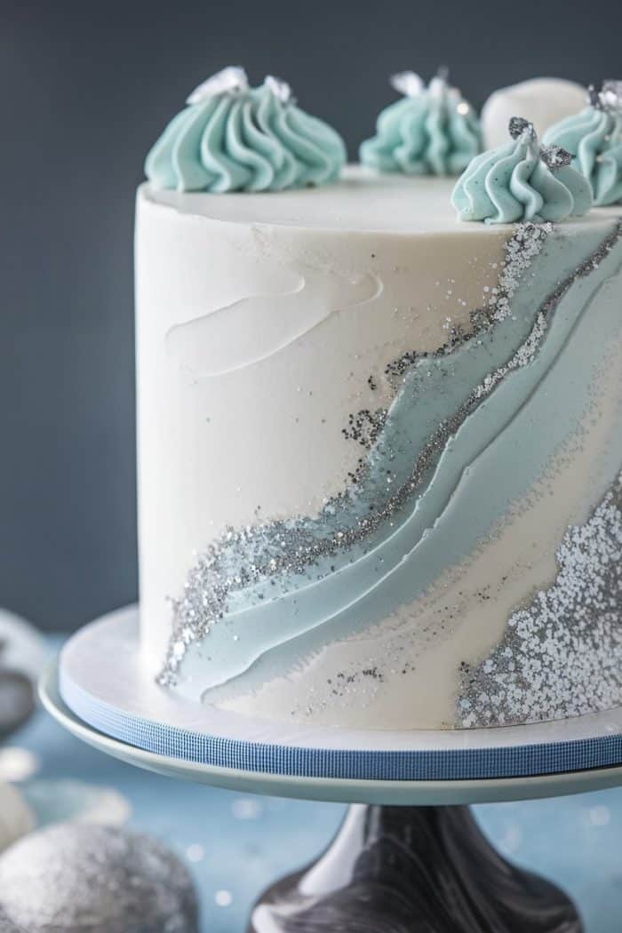 A tall, elegant cake with smooth white icing and diagonal swirls of light blue frosting. Sparkling silver edible glitter decorates the surface, with rosettes of blue frosting on top. Placed on a blue-rimmed cake stand.