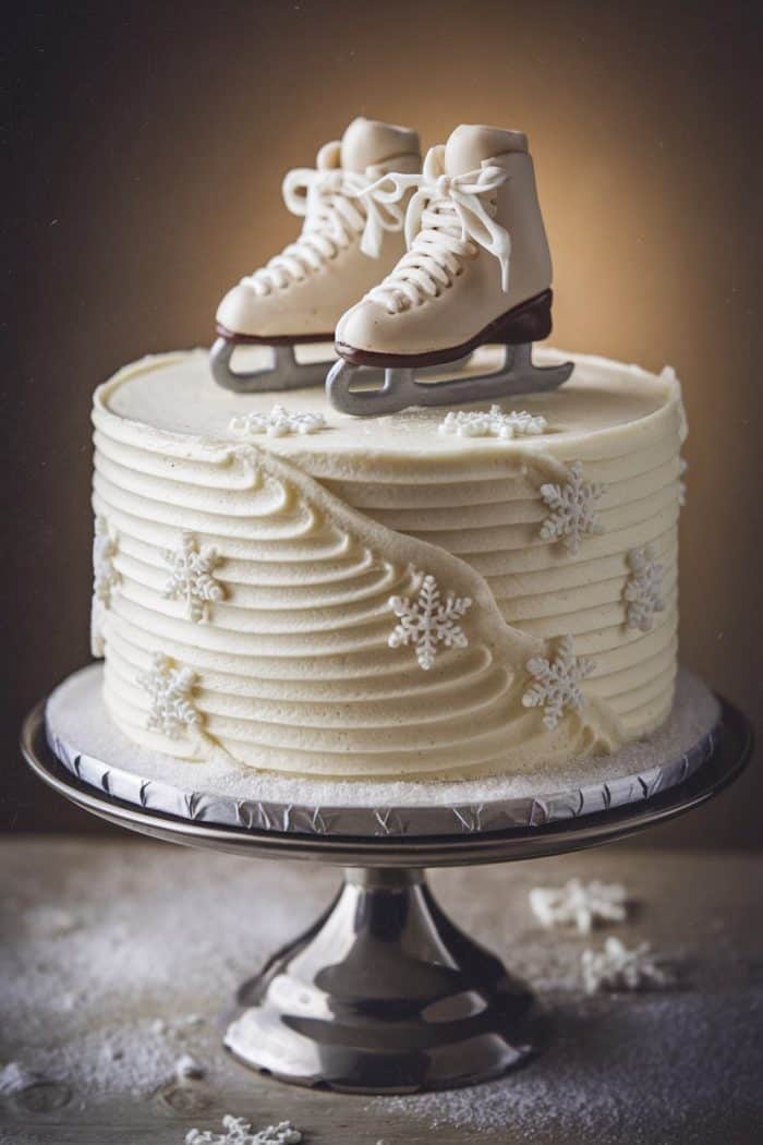 A frosted cake on a stand, decorated with delicate white frosting snowflakes. Atop the cake sits a pair of realistic-looking edible ice skates, with the background softly lit in a warm tone.