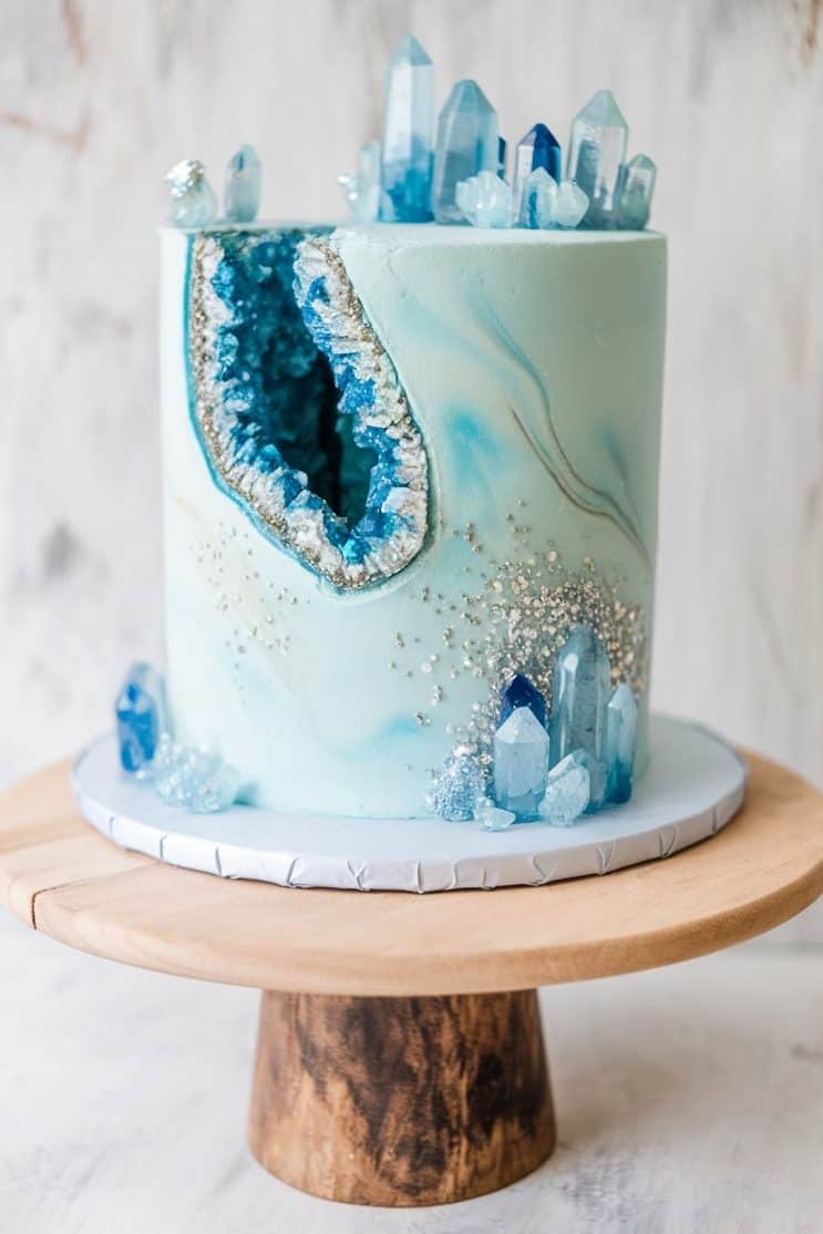 A marbled blue cake on a wooden stand features a geode design with blue crystals and silver accents. The top is adorned with clear and blue sugar crystals, creating a striking geode effect.