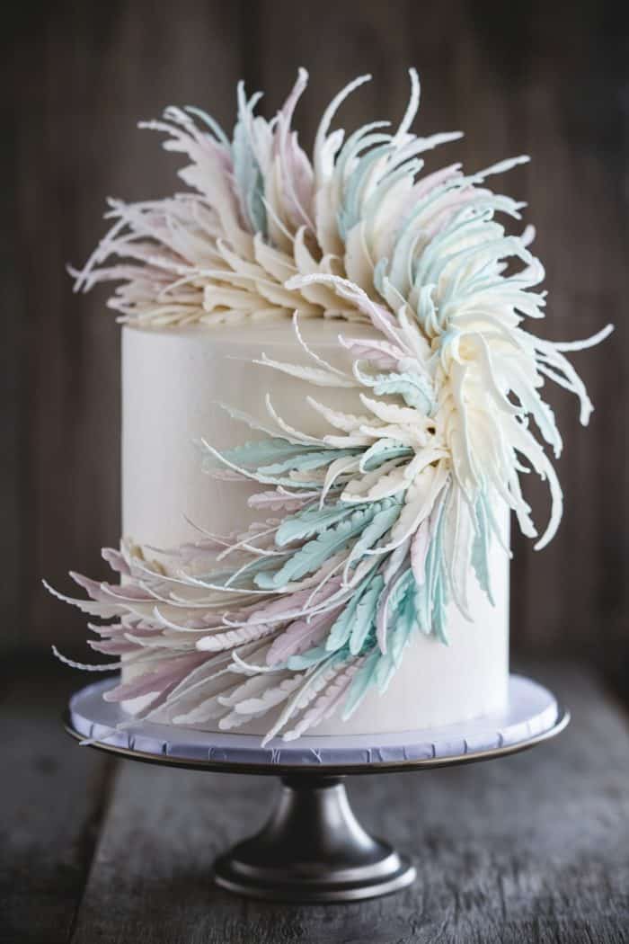 A white frosted cake adorned with intricate, pastel-colored sugar decorations resembling feathers or petals, sits elegantly on a silver cake stand. The soft hues include shades of pink, blue, and cream, creating a delicate, flowing pattern.
