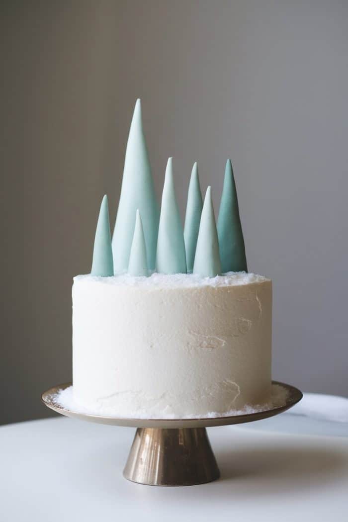 A white frosted cake on a gold stand, decorated with tall, pointed, blue and teal peaks resembling ice or mountains. The cake sits on a white table against a neutral background, creating a minimalist, elegant appearance.