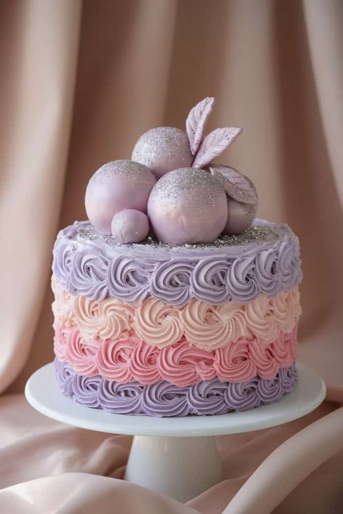 A beautifully decorated cake with layers of pink and purple swirled frosting sits on a white cake stand. It is topped with glittery, spherical decorations and leaves, all in pastel hues, set against a soft, draped fabric background.