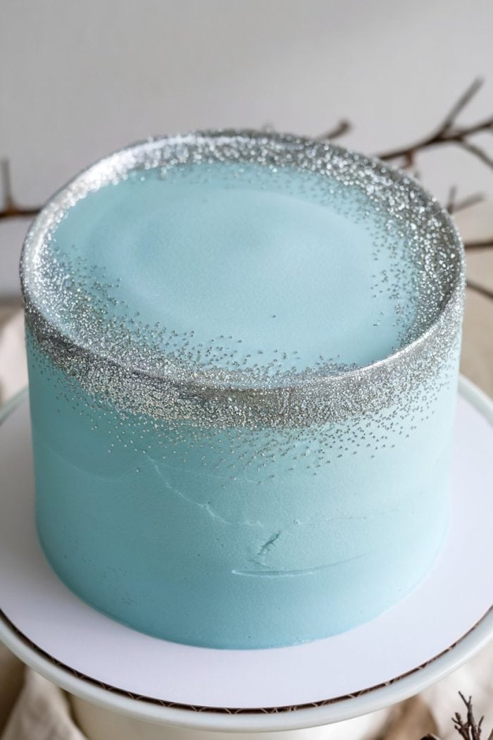 A light blue frosted cake with silver glitter sprinkled around the top edge, sitting on a white cake stand. The background features blurred branches for a simple, elegant look.