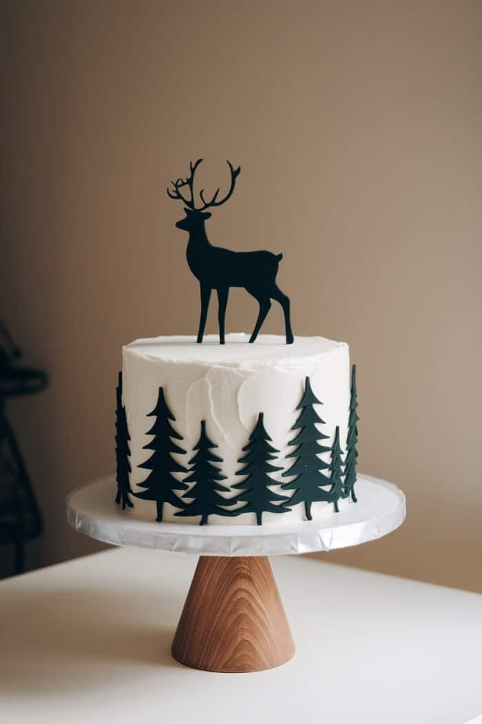 A white frosted cake on a wooden stand, decorated with black silhouettes of pine trees around the sides. A black silhouette of a deer with antlers stands on top. The cake is set against a plain background.