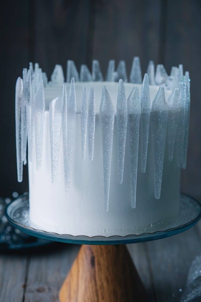 A white frosted cake adorned with clear, glittery icicle-like decorations on top, displayed on a wooden pedestal cake stand against a dark background.