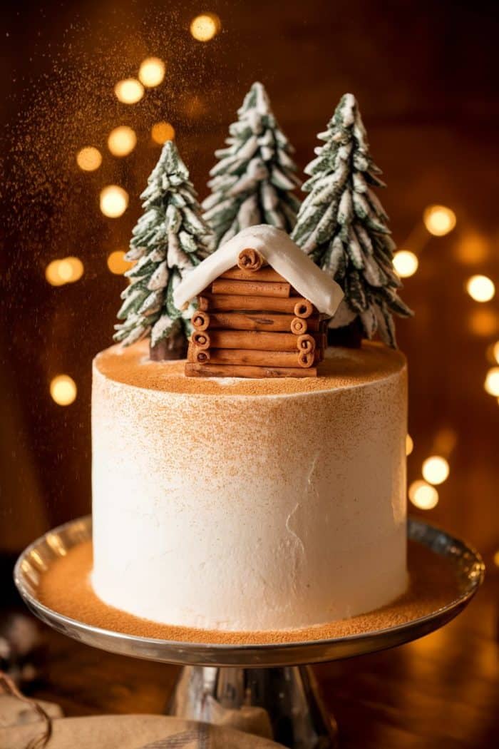A rustic cake with white frosting features a gingerbread cabin and three snow-covered pine trees on top. The cake is dusted with cinnamon, and warm bokeh lights glow in the background, creating a cozy atmosphere.
