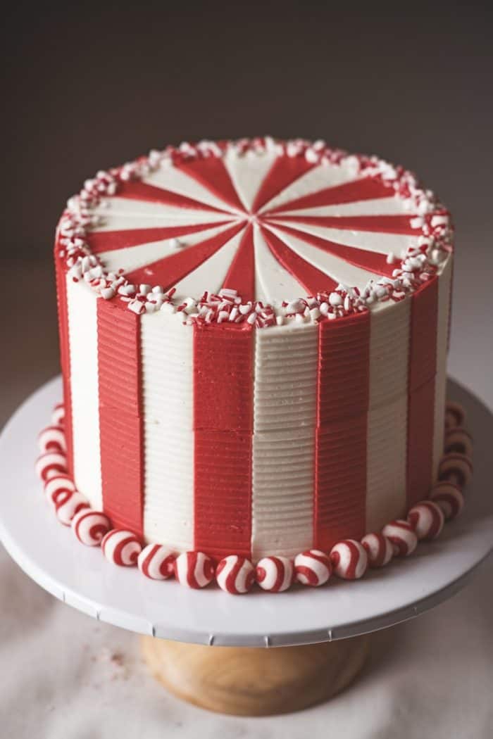A red and white striped cake with a peppermint pattern on top. The cake is decorated with peppermint candies around the base and edge. It sits on a white cake stand with a wooden base against a neutral background.