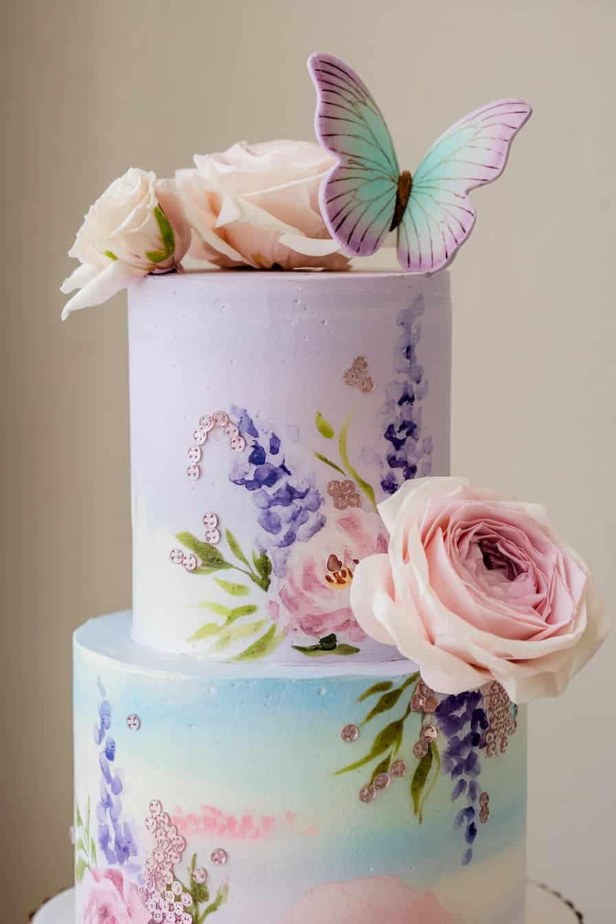 A two-tiered pastel watercolor cake adorned with large pink roses and a purple-and-teal butterfly topper. The cake features delicate floral designs with pink and purple flowers and subtle green leaves.