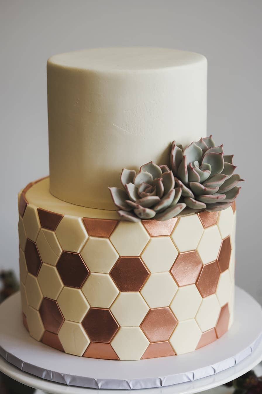A two-tiered cake with a smooth top tier and a hexagonal pattern on the bottom in white, gold, and bronze. Two realistic fondant succulents adorn the side.