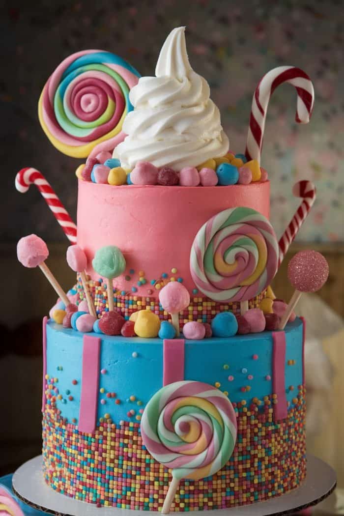 A vibrant, two-tiered cake is decorated with colorful lollipops, candy canes, and sprinkles. The bottom layer is blue with multicolored sprinkles, and the top is pink with large lollipops and marshmallows, topped with a swirl of white frosting.