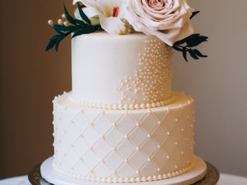 A two-tiered white wedding cake with pearl decorations and quilted patterns. Topped with a large pink rose, a white flower, and green leaves, placed on an ornate gold stand.