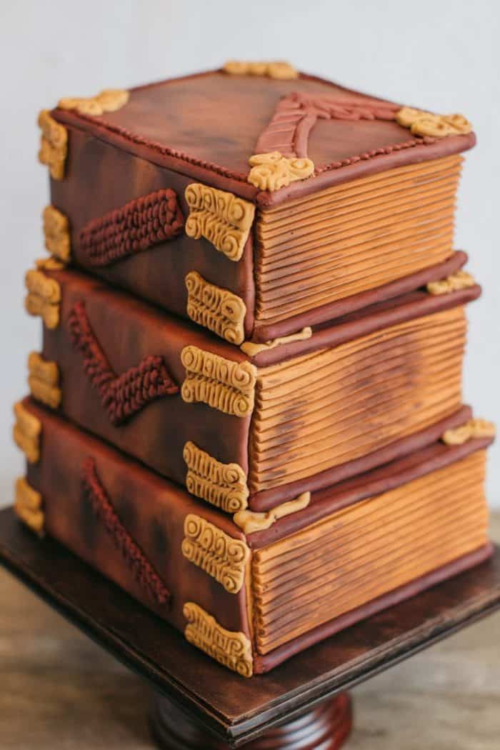 A cake designed to look like a stack of three leather-bound books with detailed edges and ornamental designs on the corners. The cake is placed on a stand, showcasing the intricate texture and color variations that mimic aged books.