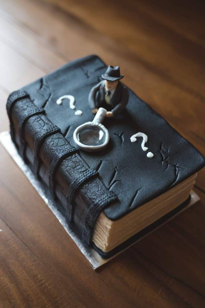 A cake designed to look like a black leather-bound book, adorned with a figure wearing a suit and hat holding a magnifying glass. White question marks are placed on the book. The cake is on a wooden surface.