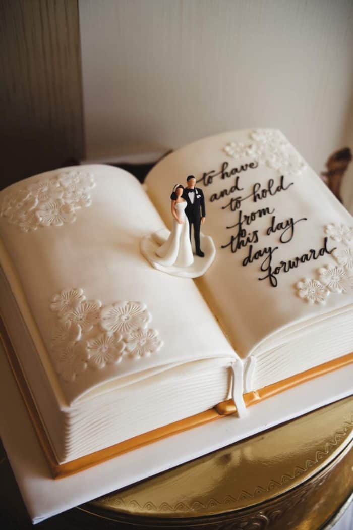A book-shaped cake with floral patterns on white icing. A figure of a bride and groom stands atop. Written on the cake: "to have and to hold from this day forward." The cake sits on a gold platter.