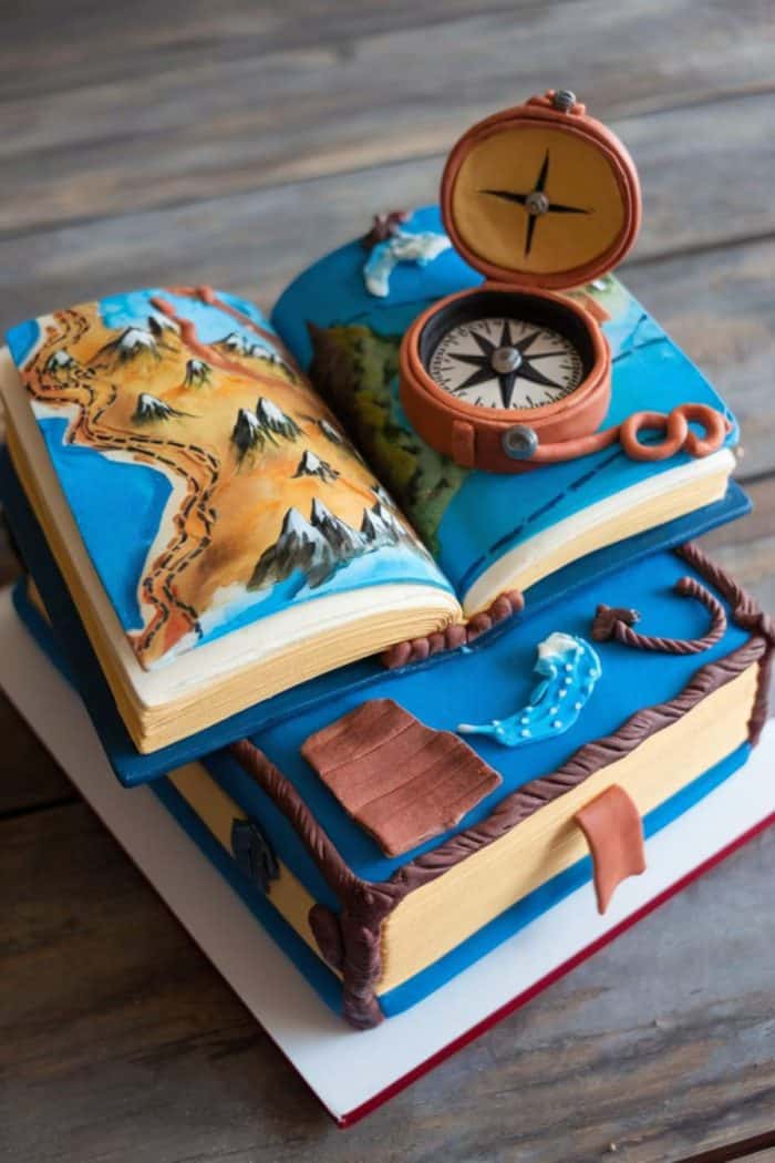 A cake shaped like two stacked books with a decorative map and compass on top. The book covers are blue with brown accents. The scene includes mountains and water features crafted from icing. The cake is placed on a wooden surface.
