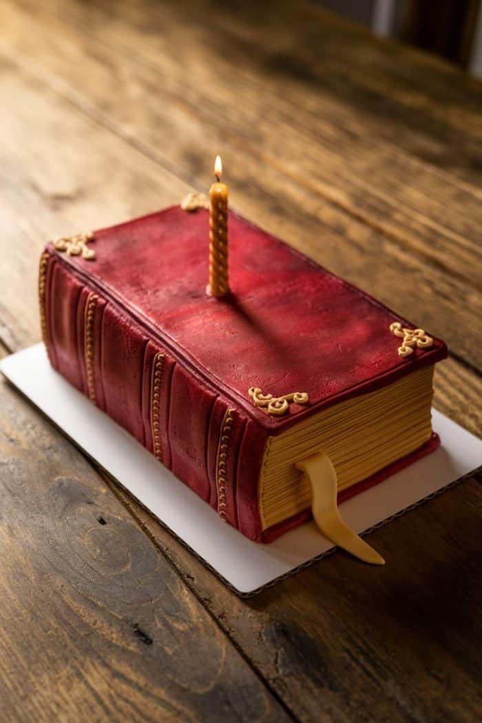 A cake designed to look like a leather-bound book with a red cover and gold accents sits on a wooden table. A single lit candle is positioned on top of the cake.