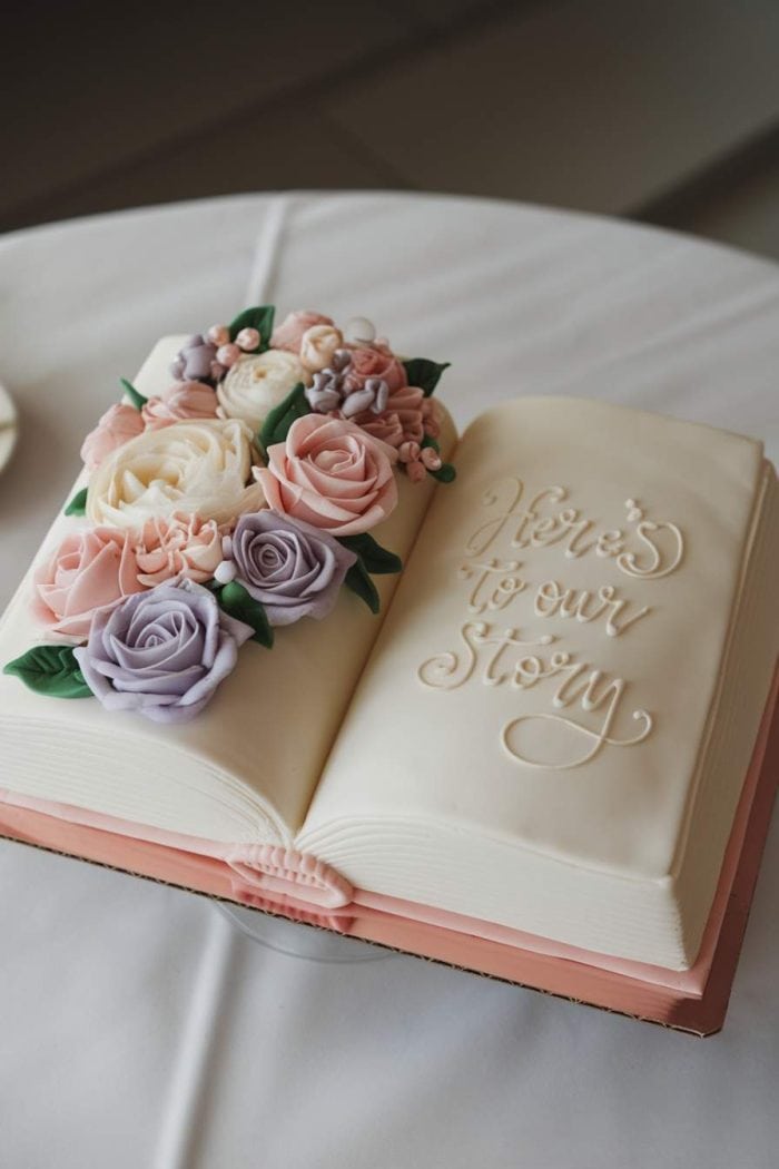 A cake designed to look like an open book with the words "Here's to our Story" written on one page. The cake is decorated with pastel roses in pink, purple, and cream, arranged along the center and one side.