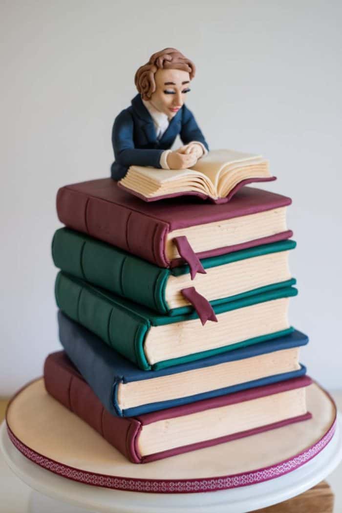 A cake designed like a stack of four books with a fondant figure of a person reading on top. The books are green, blue, and maroon, with realistic texturing and ribbons as bookmarks. The cake sits on a round, decorated base.