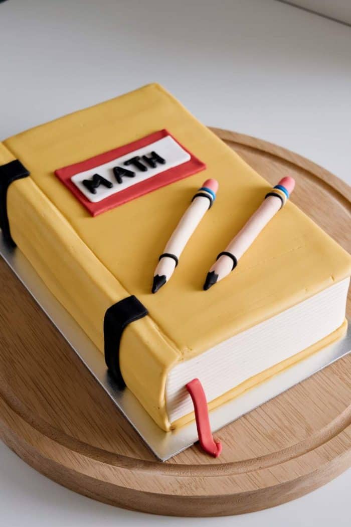 A cake shaped like a closed yellow math book rests on a wooden platter. It features fondant details, including two pencils and a red bookmark. The word "Math" is displayed on the cover.