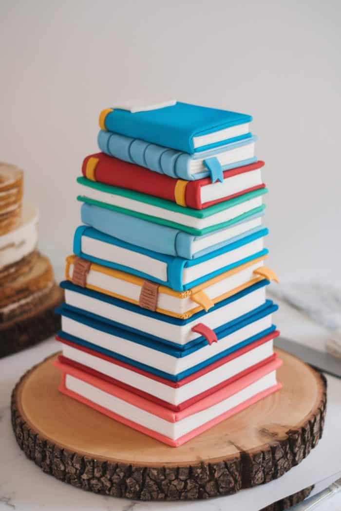 A cake designed to look like a stack of colorful books, with detailed bindings and ribbon bookmarks, placed on a wooden slab.