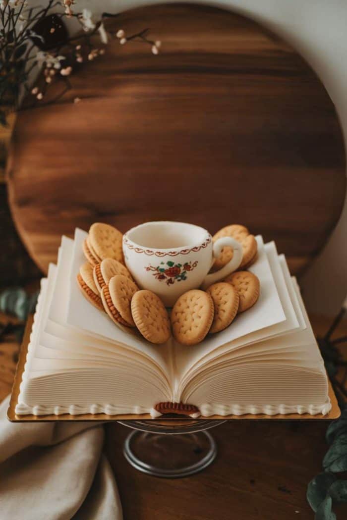 A cake designed to look like an open book with a floral teacup filled with tea on top. The cake is topped with round, embossed cookies arranged around the cup. A wooden board is in the background.