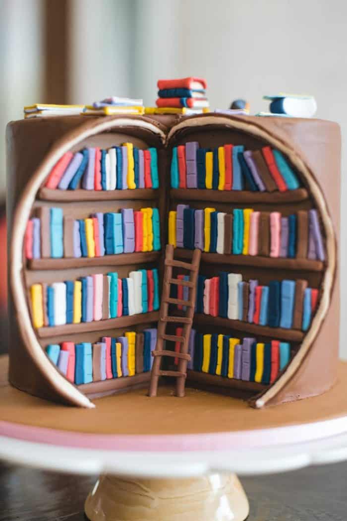 A round cake designed as a brown leather-bound book, with the front cut open to reveal edible books on shelves inside. A small ladder leans against the shelves, and a few decorative books rest on the cake's top.