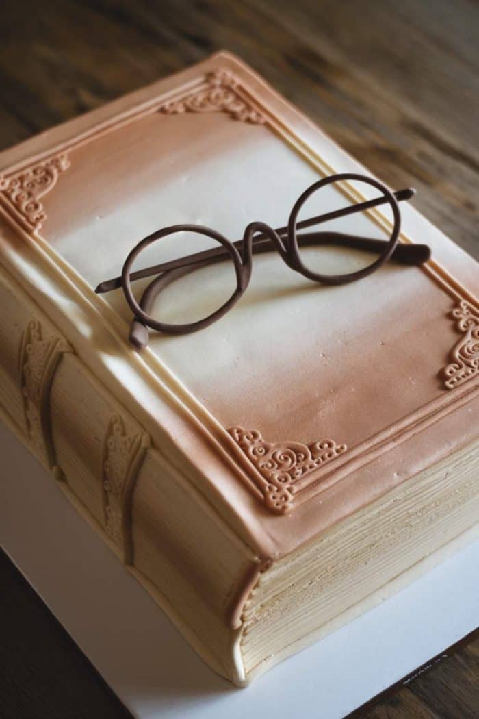 A cake designed to look like an ornate, leather-bound book with realistic pages, featuring a pair of chocolate glasses on top. The cake rests on a wooden surface.