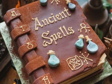 A cake designed to look like an ancient spellbook, featuring intricate chocolate detailing, edible potion bottles, and mystical symbols. The cover reads "Ancient Spells" in golden letters. The cake is set on a wooden table.
