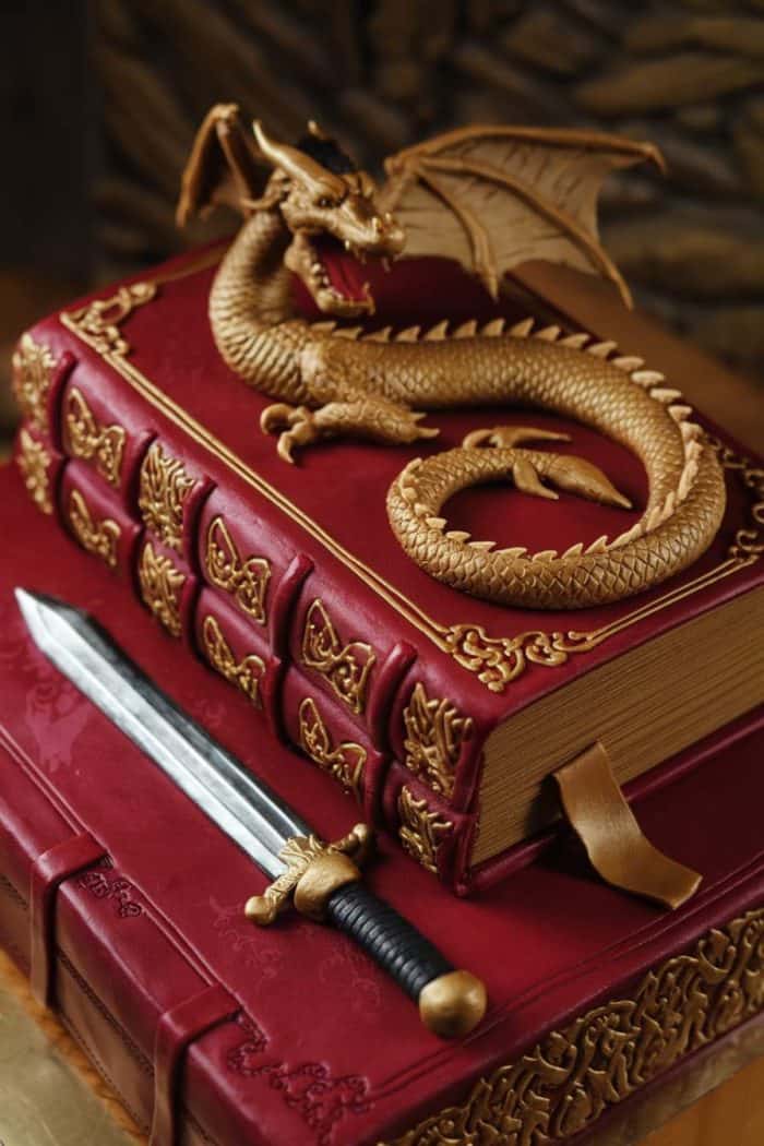 A decorative cake designed as two leather-bound books with ornate gold detailing. A gold dragon sits on top, and a realistic sword with a black handle rests alongside. The cake features intricate craftsmanship.