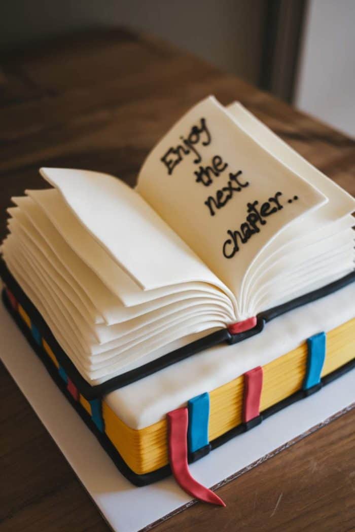 A cake designed to look like an open book with realistic pages. The top page is inscribed with "Enjoy the next chapter..." in black icing. The cake layers resemble colorful book edges, with ribbons cascading down the sides.